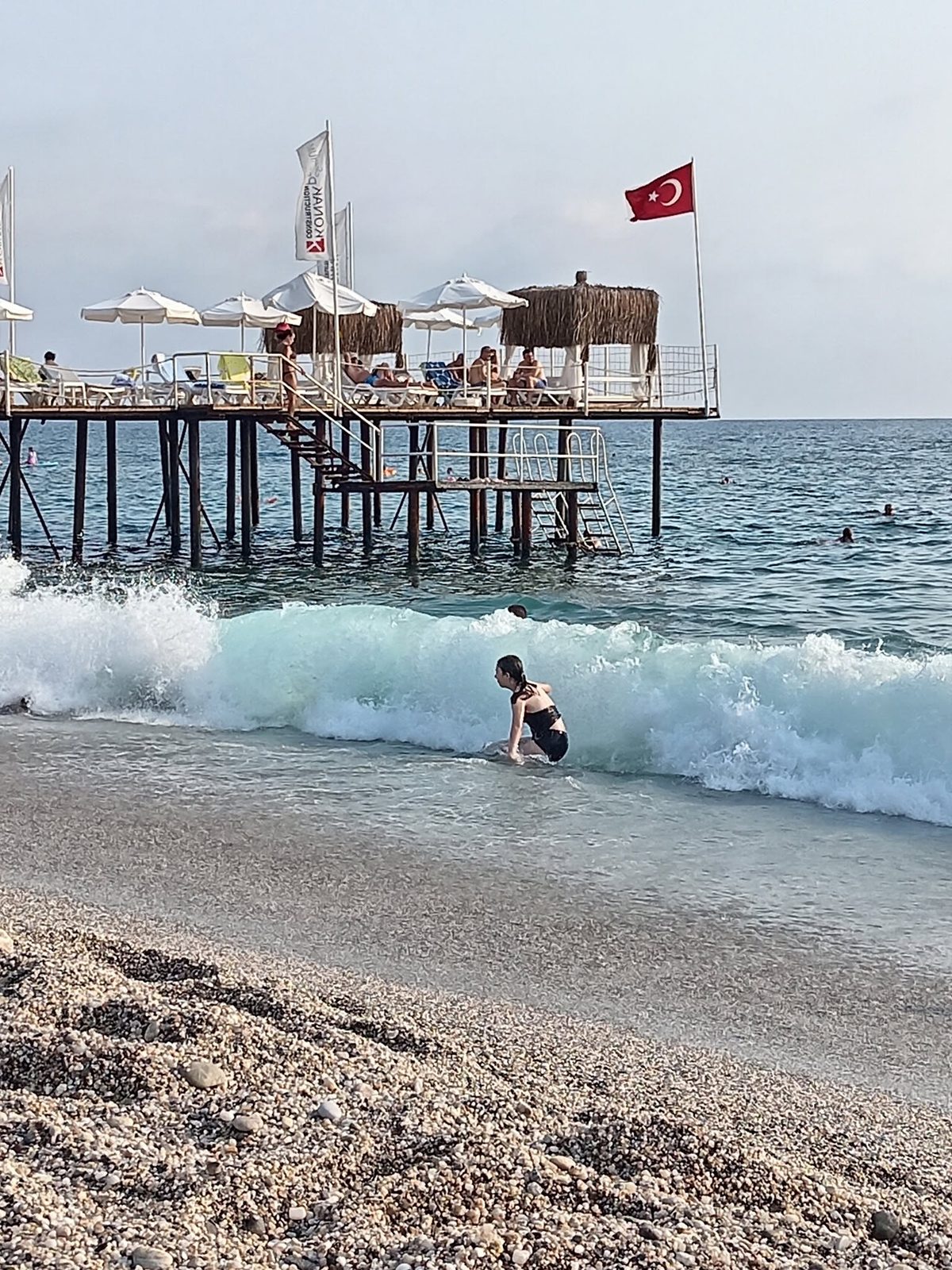 A beach with waves and a wooden pier. A flag flies on the pole.